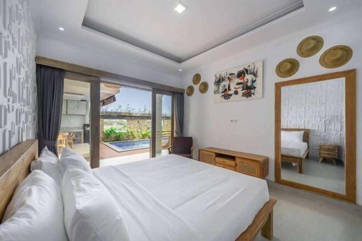 Une chambre moderne avec un grand lit, une tête de lit en bois, un bureau et une chaise. Une porte-fenêtre coulissante donne accès à un balcon avec vue sur la verdure.