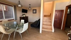 Een eethoek met een glazen tafel en witte stoelen, een houten trap en een televisie aan de muur.