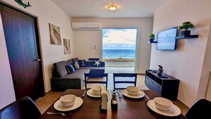 A compact living space with a dining area, a sofa facing a large window overlooking the sea, and a television mounted on the wall. - Stylish Ocean view w/ HotTub BBQ (Arecibo)