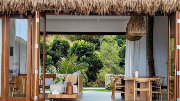A modern outdoor living area with a thatched roof, a wooden dining set, and a sofa with cushions. - Umarato Villa Sumba (Wanokaka)