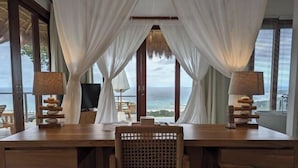 A wooden desk with two lamps, a woven chair, and a view of the ocean through sheer curtains. - Umarato Villa Sumba (Wanokaka)