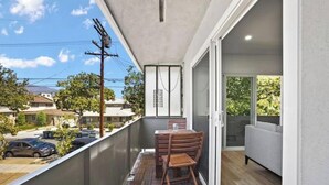 Een balkon met een houten tafel en stoelen, een grijze bank en uitzicht op de straat en huizen.