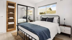 Une chambre à coucher comprenant un lit, une armoire, une table de chevet, une lampe et une fenêtre donnant sur des immeubles.