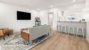 A modern living room with a sofa, a coffee table, and a TV mounted on the wall. There is a kitchen area with a counter and stools. - Spacious Apt w/1BR - City Center (Pasadena)