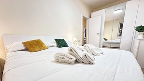 Een net opgemaakt bed met witte lakens en opgevouwen handdoeken, een geel-groen kussen en een witte kledingkast met spiegel.