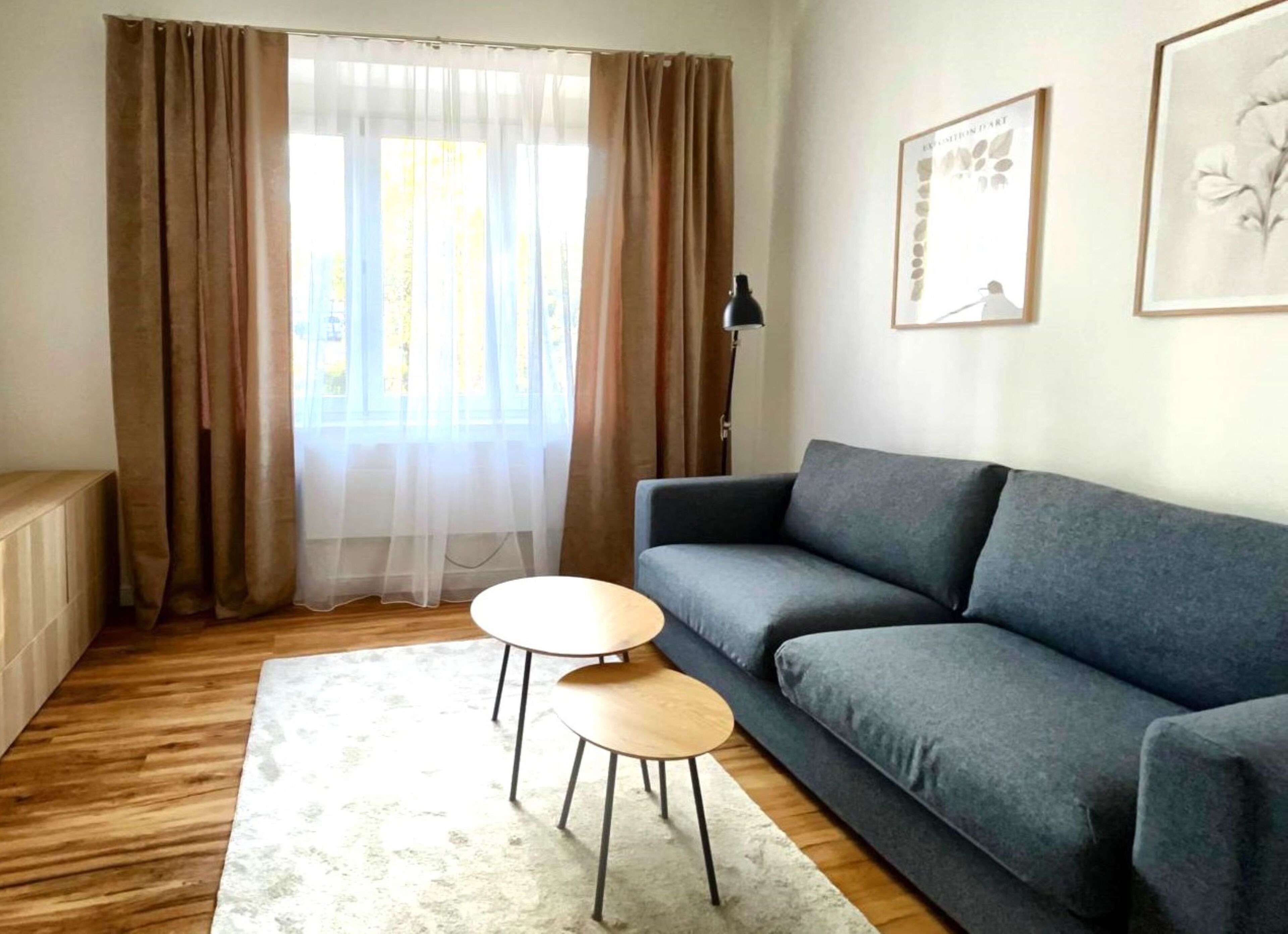 A living room with a grey sofa, two wooden side tables, a white rug, and a window with sheer curtains.