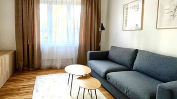 A living room with a grey sofa, two wooden side tables, a white rug, and a window with sheer curtains.