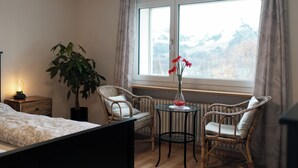 A bedroom with a bed, a nightstand, a potted plant, two wicker chairs, a small round table with a vase of flowers, and a window with a view of mountains. - Epic view and sun sets from spacious 4 bedroom apartment. Large families +groups (Amden)