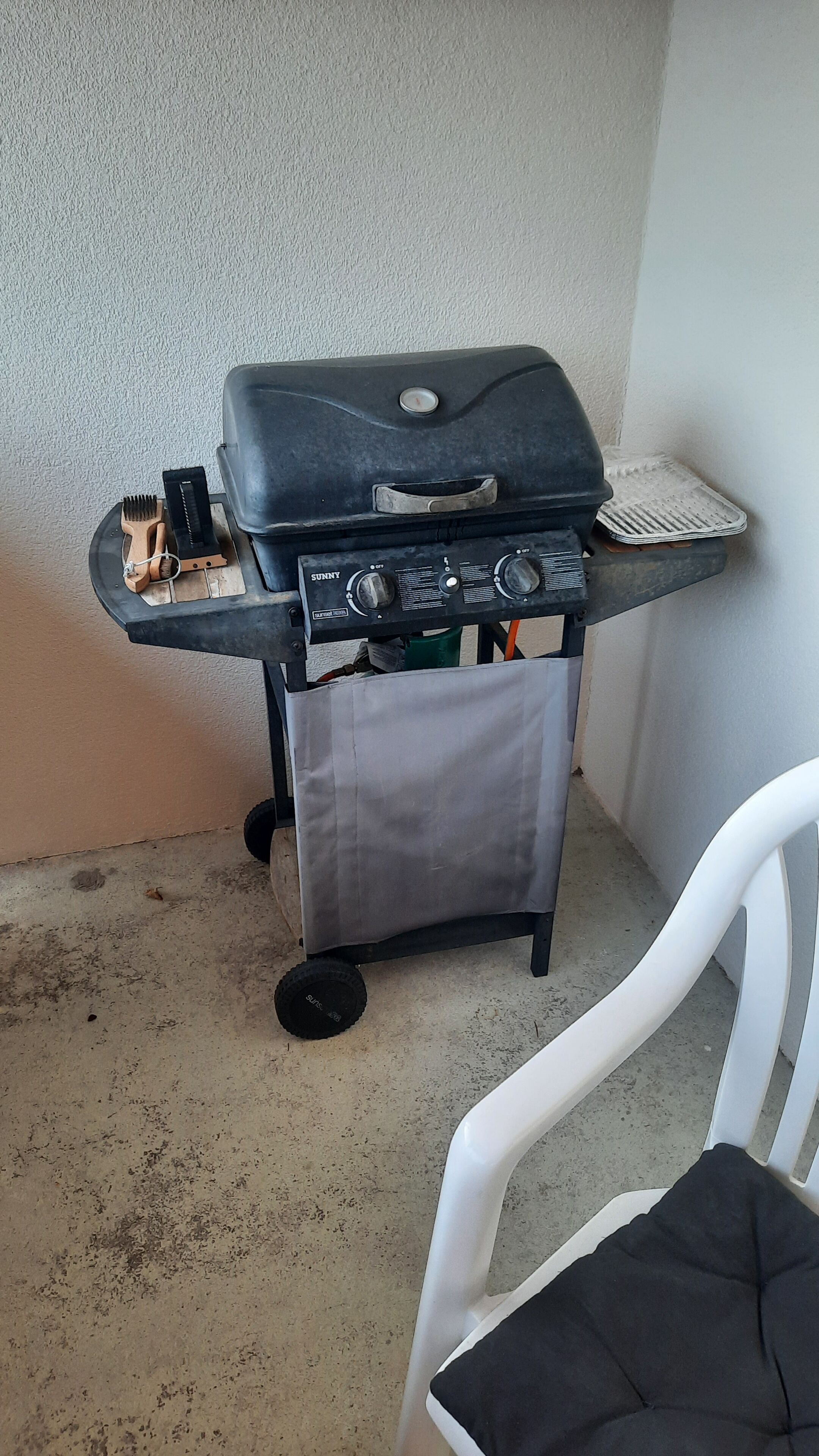 A portable grill with controls and a side burner, placed on a small cart with wheels, against a wall.