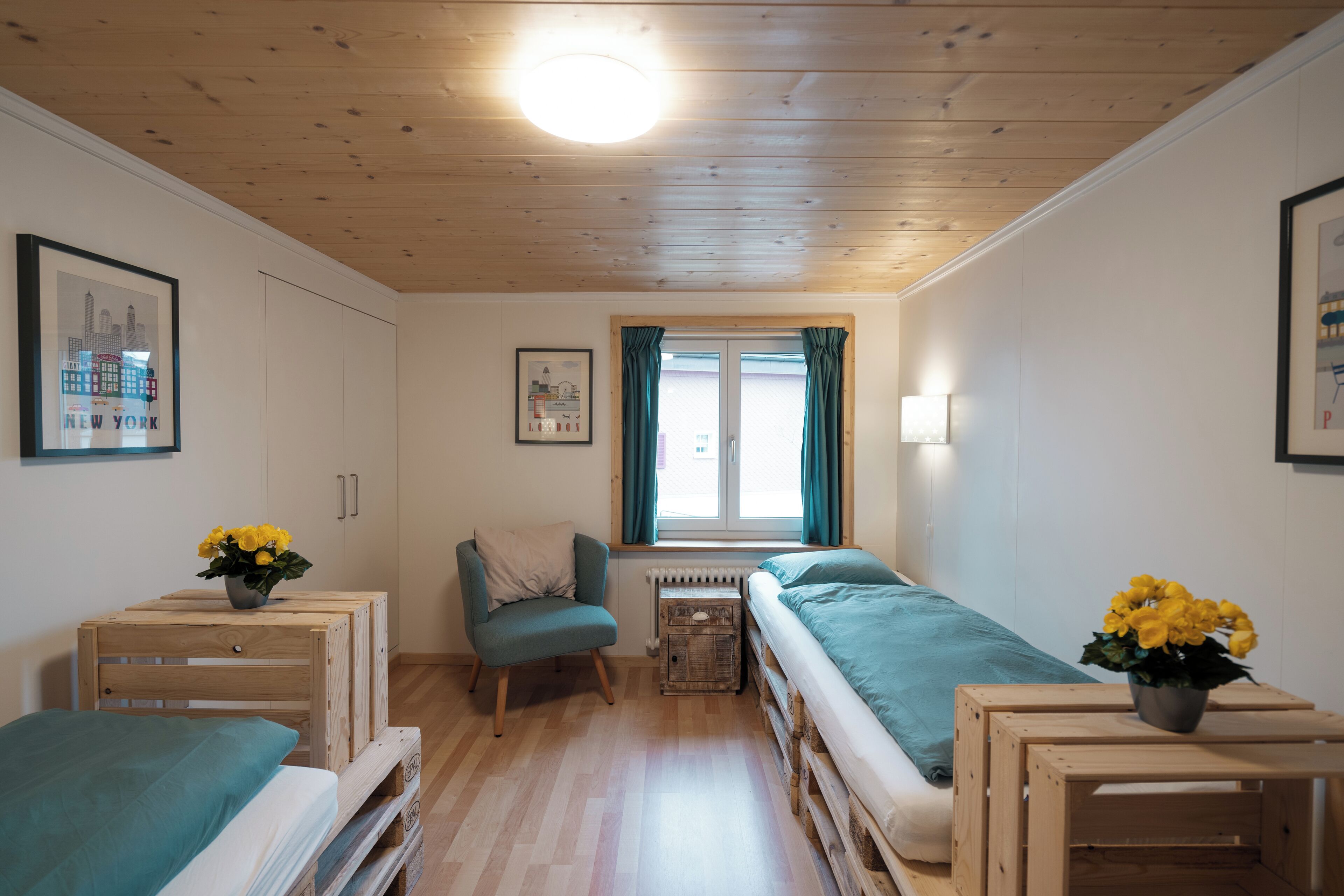A small, cozy room with a wooden ceiling, a single window with curtains, a bed, a chair, and a small table with flowers.