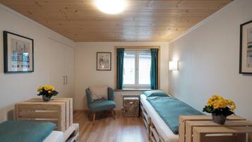 A small, cozy room with a wooden ceiling, a single window with curtains, a bed, a chair, and a small table with flowers.
