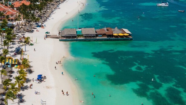 Vista aérea de una playa con un edificio grande de varios techos, probablemente un resort u hotel, junto a la costa.