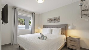Un dormitorio con una cama grande, una mesita de noche, un televisor montado en la pared y una ventana con cortinas.