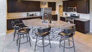 Une cuisine moderne avec des meubles en bois foncé, un comptoir en granit, un réfrigérateur en acier inoxydable, un four encastré et quatre tabourets de bar.