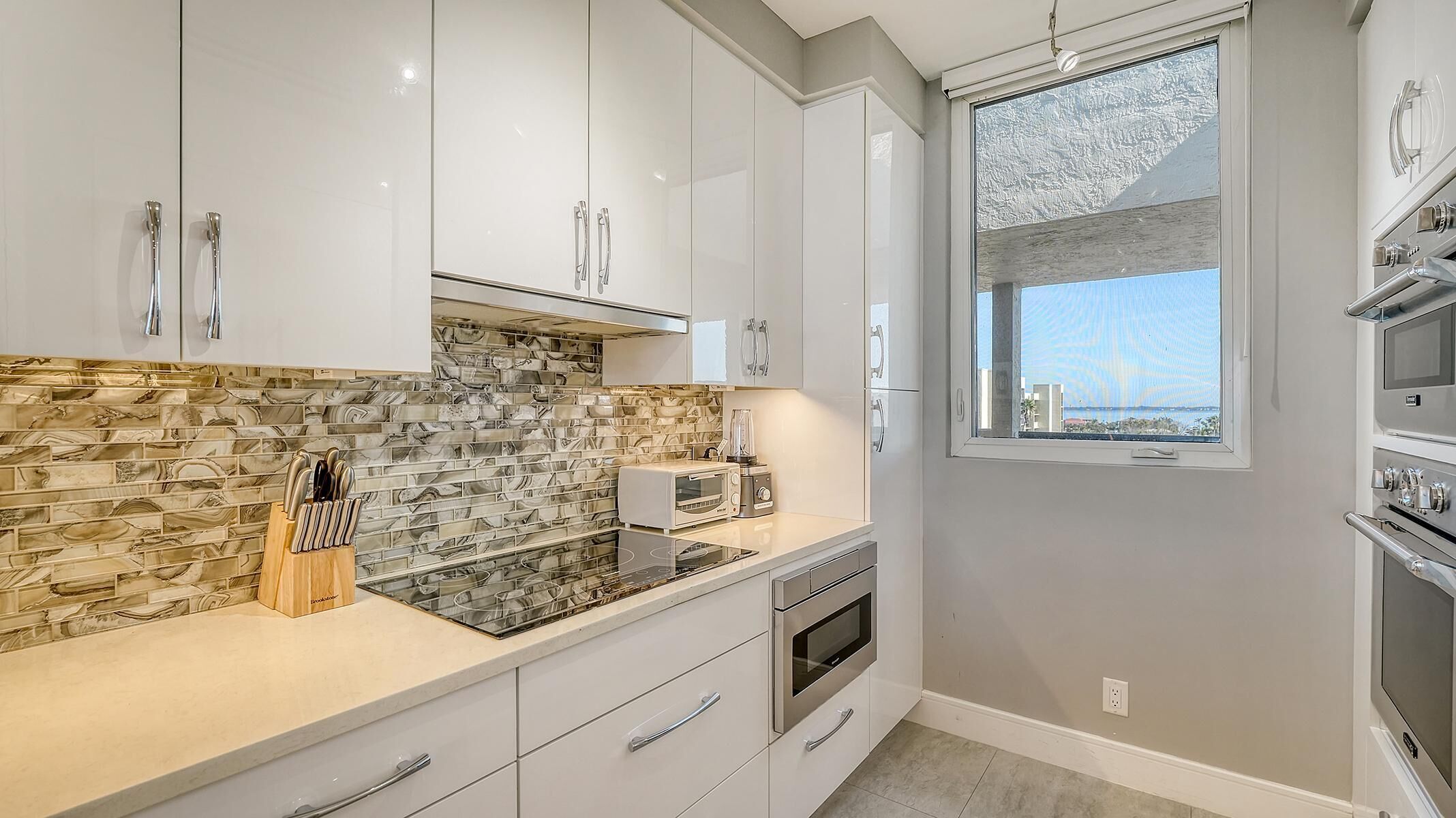 A modern kitchen with white cabinets, a marble backsplash, a built-in oven, and a window with a view.