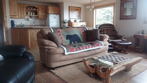 Ein gemütliches Wohnzimmer mit einer Couch, einem Couchtisch und Blick auf eine Küche mit Holzschränken und einem Kühlschrank.