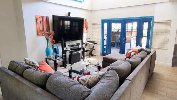 Een woonkamer met een grijze hoekbank, een flatscreen televisie, een glazen salontafel en een blauwe deur met ramen.