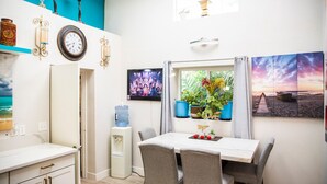 Een gezellig, compact ingerichte ruimte met een eethoek, een waterdispenser, een groot raam met planten en een televisie aan de muur.