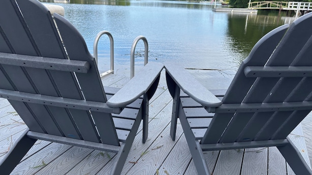 Dos sillas Adirondack mirando a un muelle de madera sobre un cuerpo de agua, con un muelle y un pequeño puente al fondo.