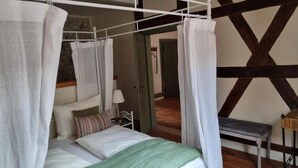 Un lit à baldaquin avec des rideaux blancs, un plaid vert et un coussin rayé. Une petite table avec une lampe se trouve à côté du lit. La pièce est dotée de poutres apparentes en bois au plafond.