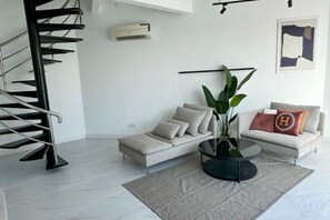 Un salon moderne comprenant un canapé, une table basse, une plante en pot, un escalier et un climatiseur fixé au mur.