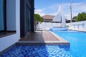 Una piscina con suelo de baldosas, un velero y un edificio blanco con techo inclinado.