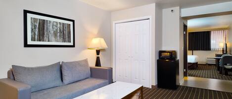 A hotel room with a gray sofa, a white coffee table, a framed picture on the wall, a door, a small table with a lamp, a microwave, and a TV.