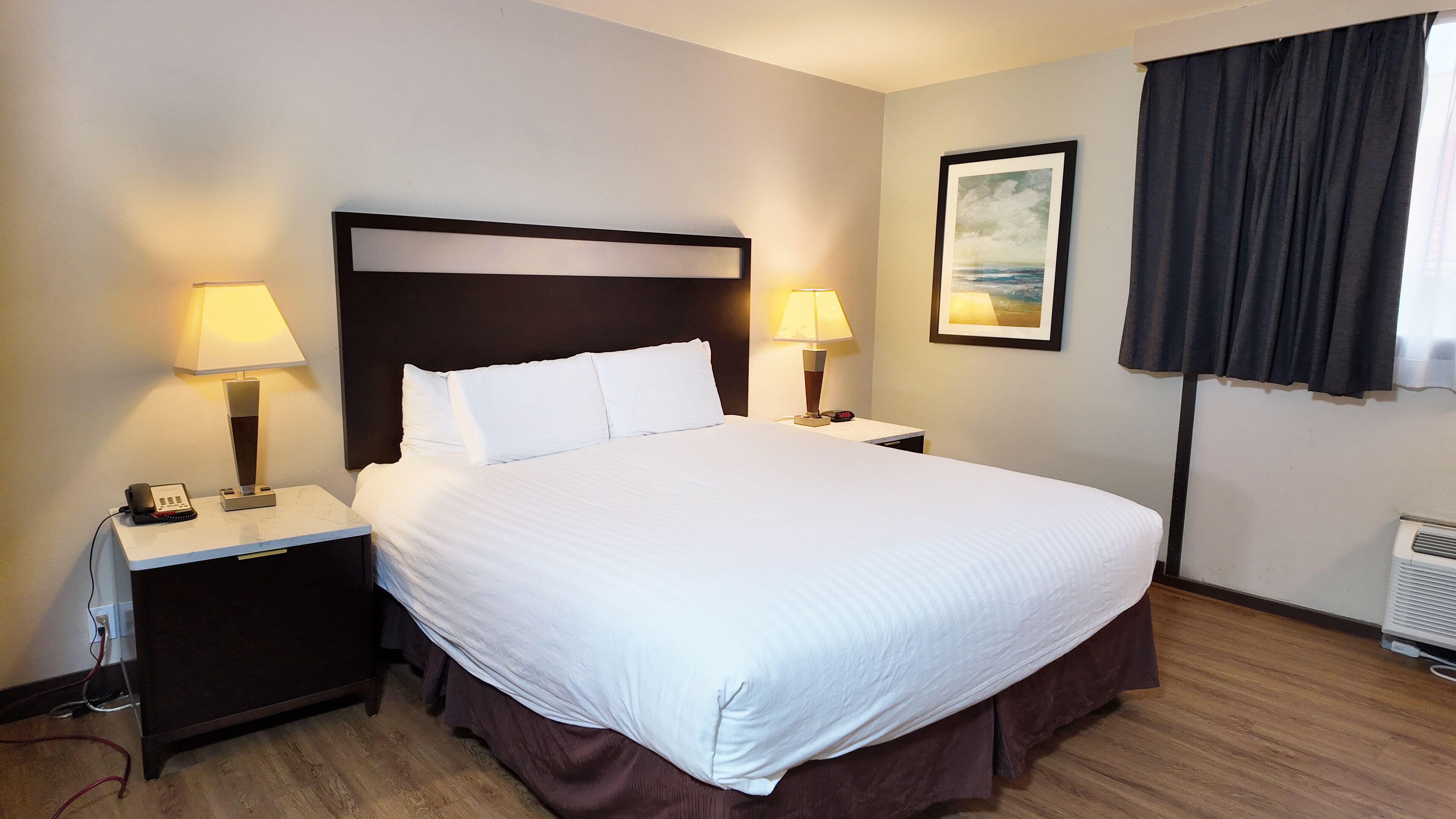 A hotel room with a large bed, two bedside tables with lamps, a painting on the wall, and a window with curtains.