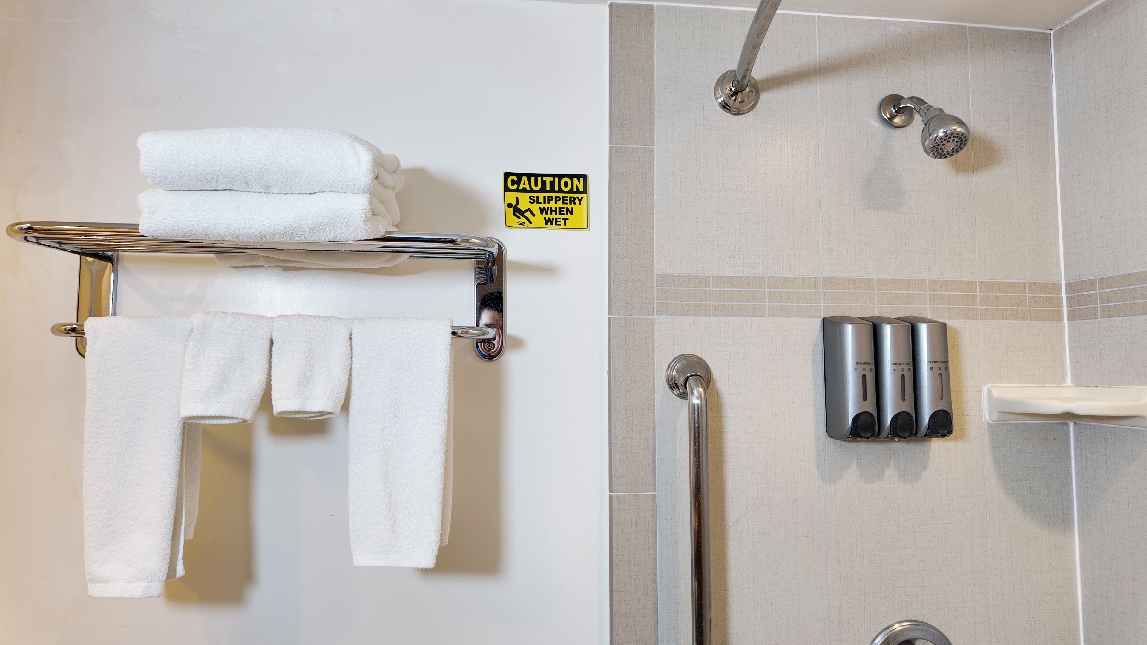 Un baño con un perchero con toallas dobladas, un letrero de precaución, un cabezal de ducha y un dispensador de jabón para manos.