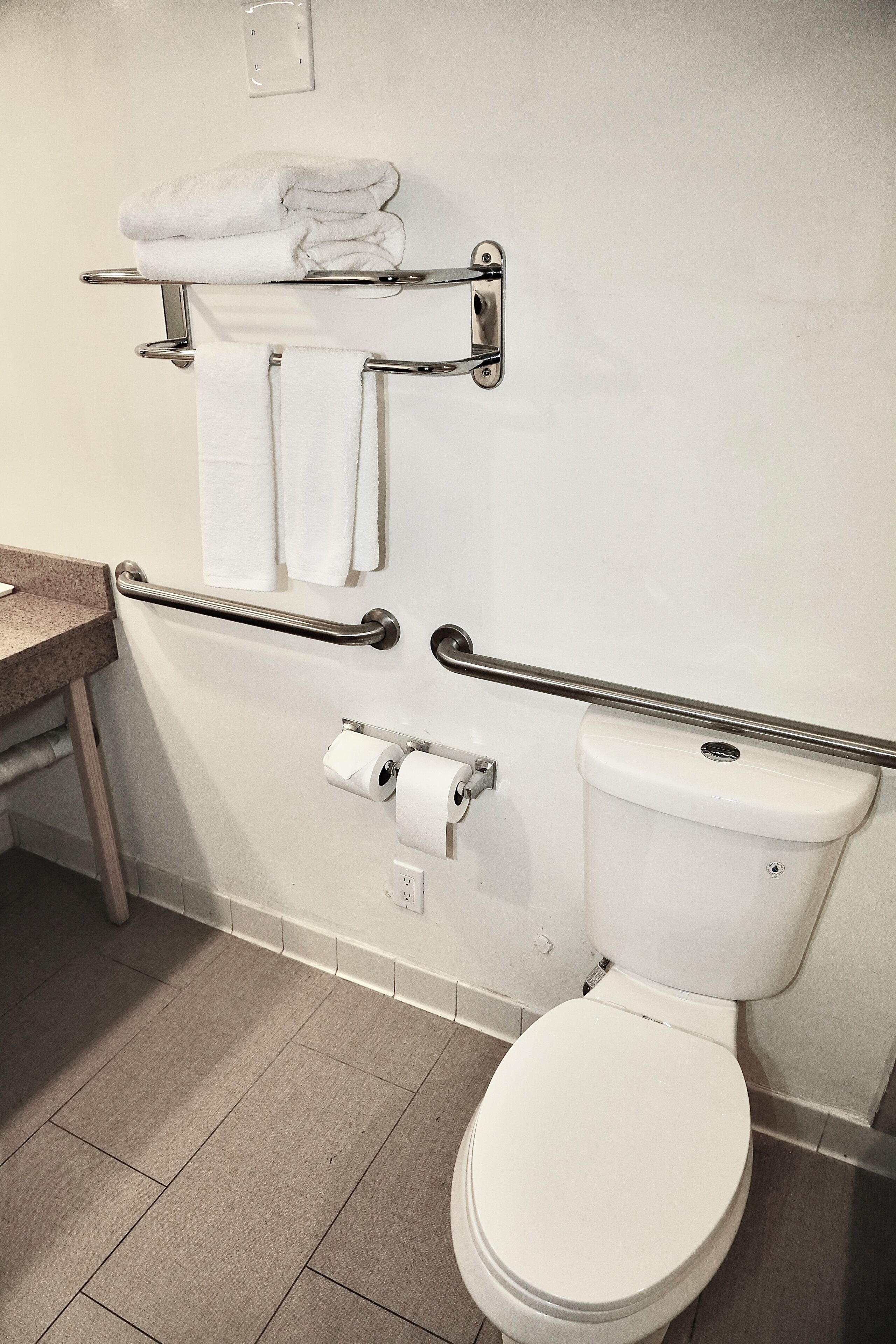 Et badeværelse med toilet, et håndklædestativ med foldede håndklæder, gelænder og toiletpapirholder.