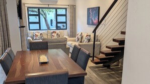A modern living room with a wooden dining table, grey upholstered chairs, a sofa with floral cushions, a staircase, and a large window with curtains.