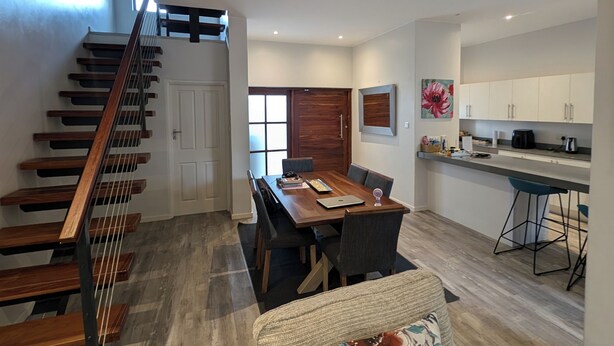 A spacious living area with a wooden staircase, a dining table with chairs, a sofa with a floral cushion, and a kitchen area with cabinets and a countertop.
