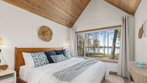 Ein Schlafzimmer mit einem großen Bett, Holzboden, Blick auf Bäume und Wasser und einem kleinen Tisch mit einer Schüssel.