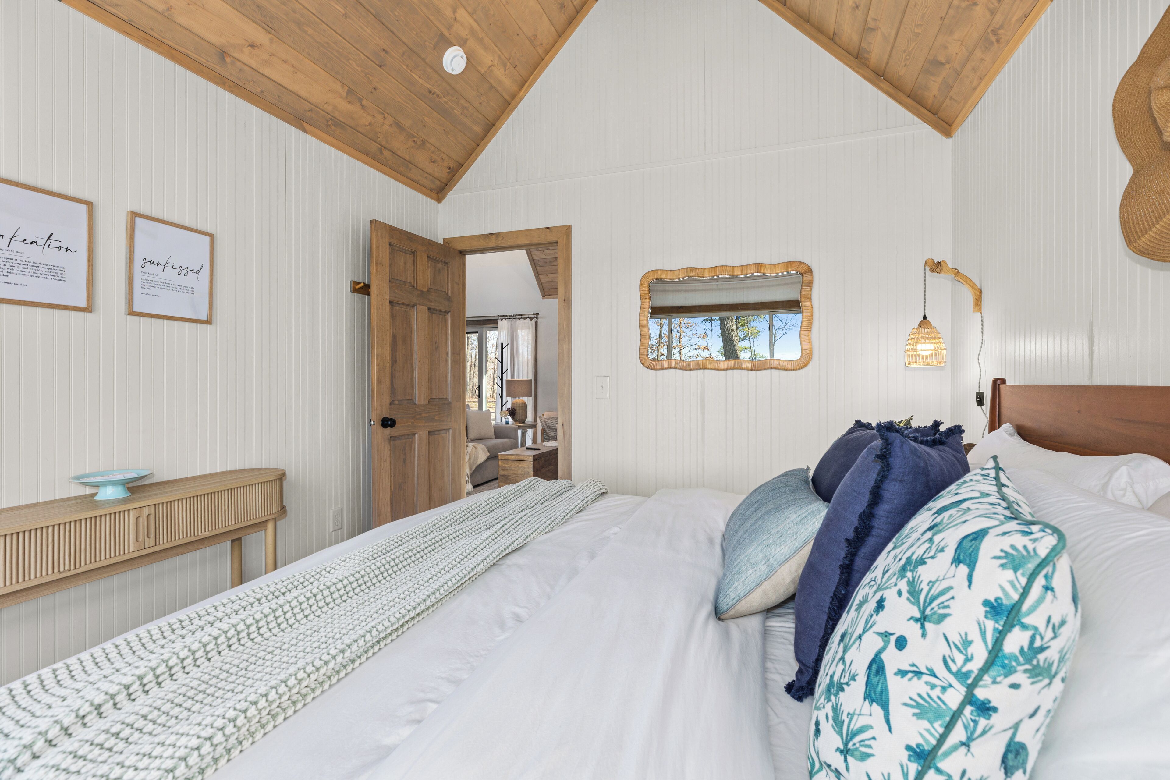 Una camera da letto con soffitto in legno, un letto con biancheria bianca, un comodino in legno, una citazione incorniciata al muro e una finestra con vista sugli alberi.