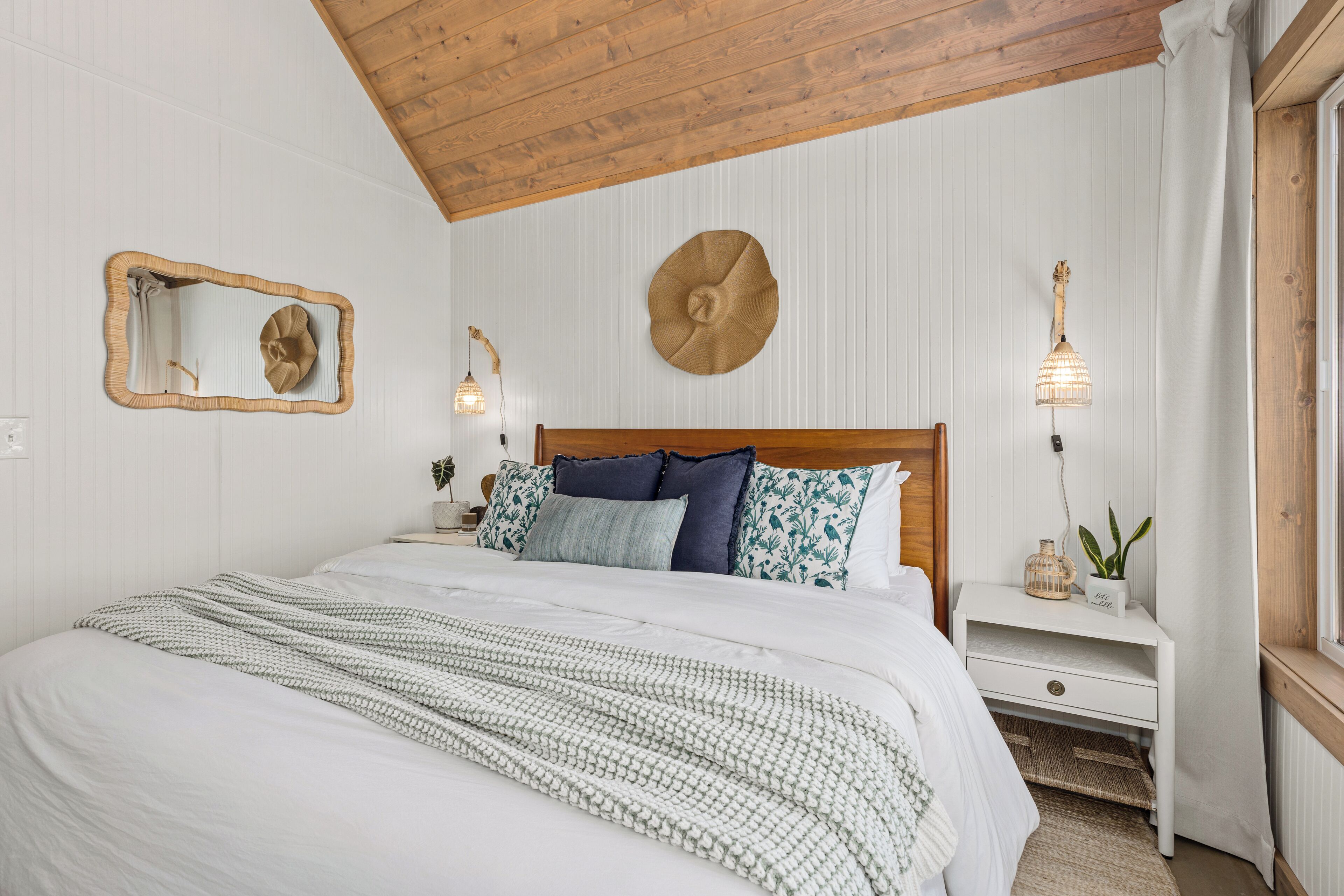 Un dormitorio con techo de madera, una cama con ropa blanca, una mesita de noche con lámpara y un espejo montado en la pared.