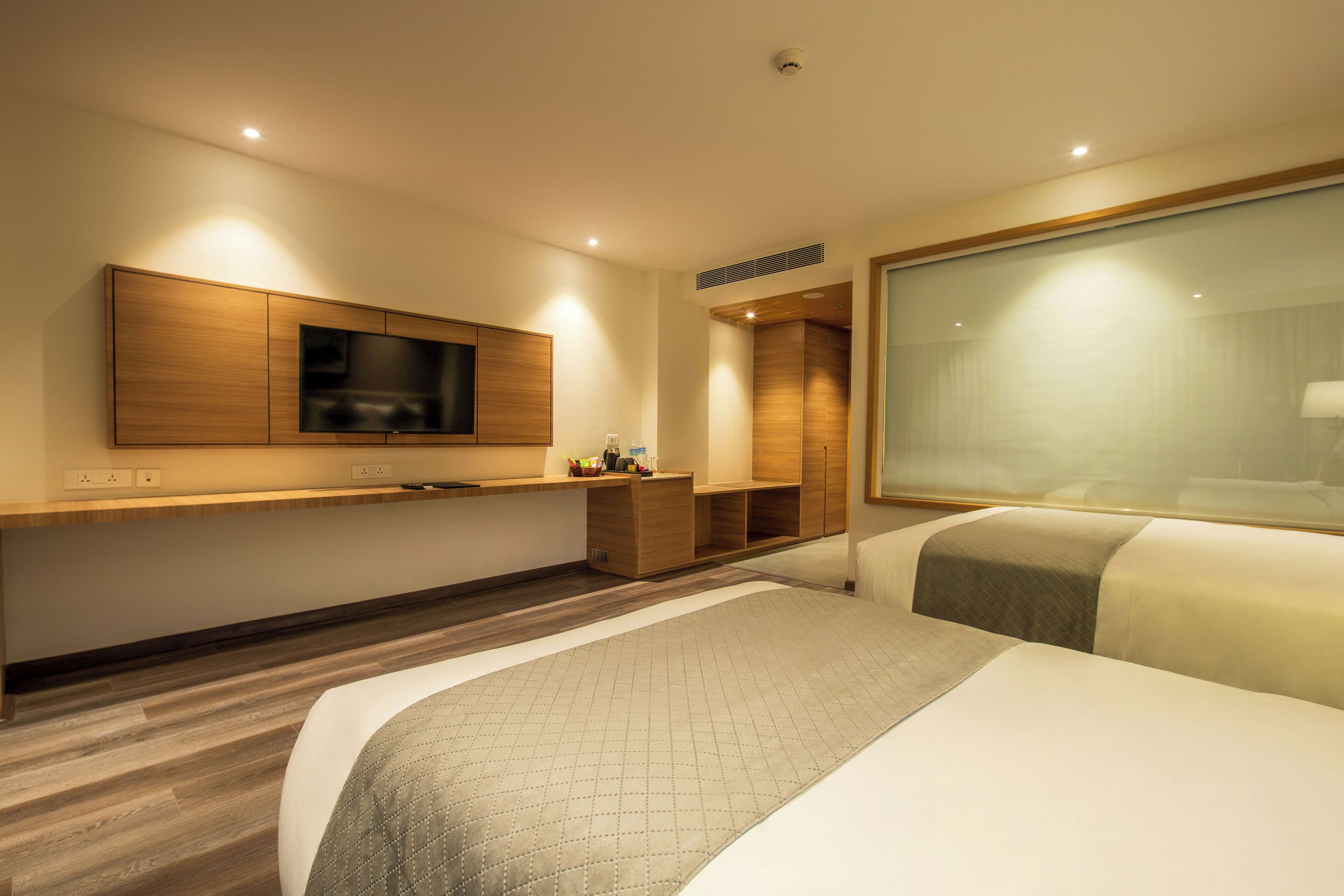 A modern hotel room with a large bed, a TV mounted on the wall, a wooden cabinet, and a large window.