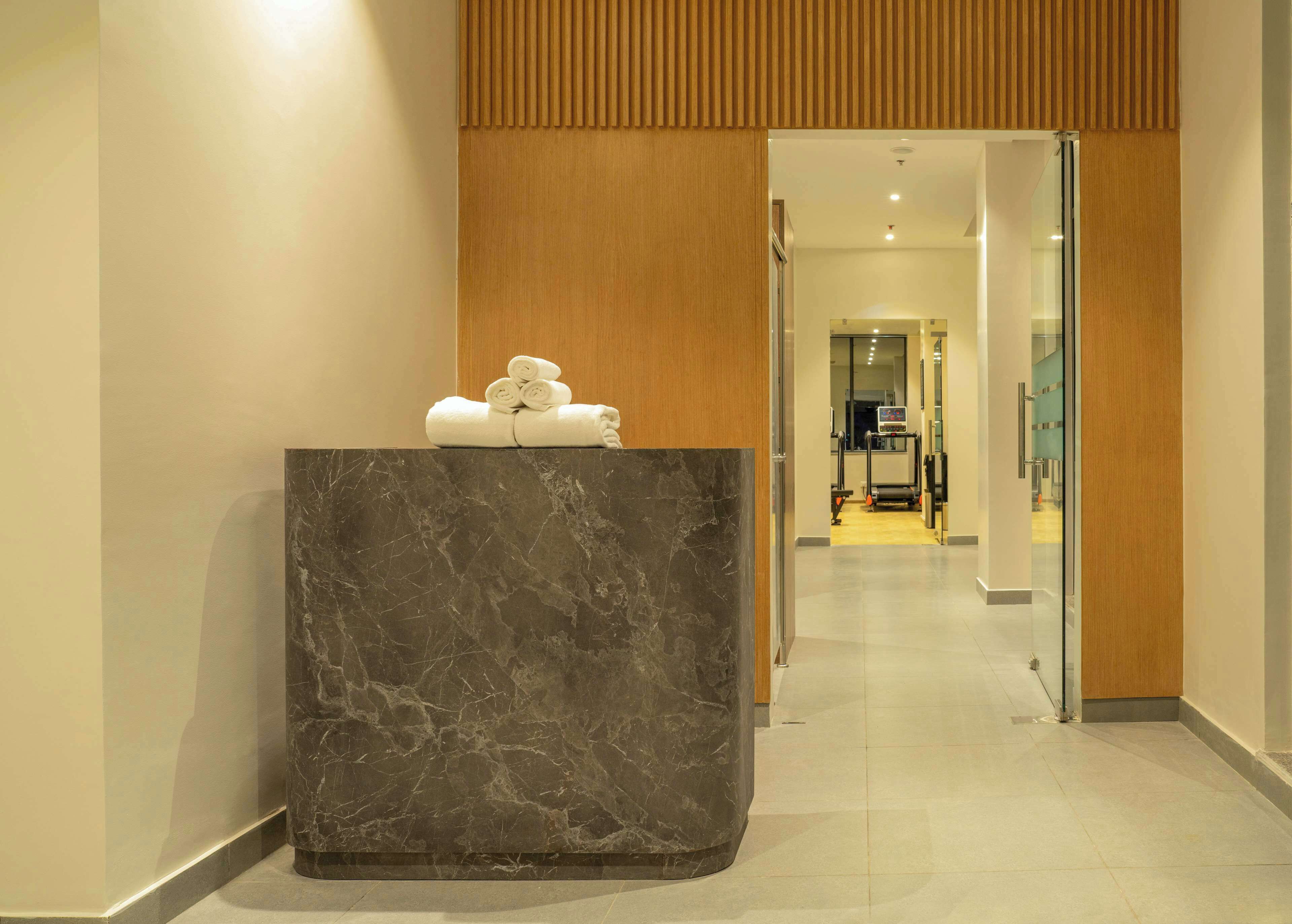 A reception area with a marble counter, a wooden panel above, and a glimpse of a fitness area in the background.