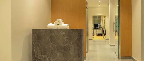 A reception area with a marble counter, a wooden panel above, and a glimpse of a fitness area in the background.