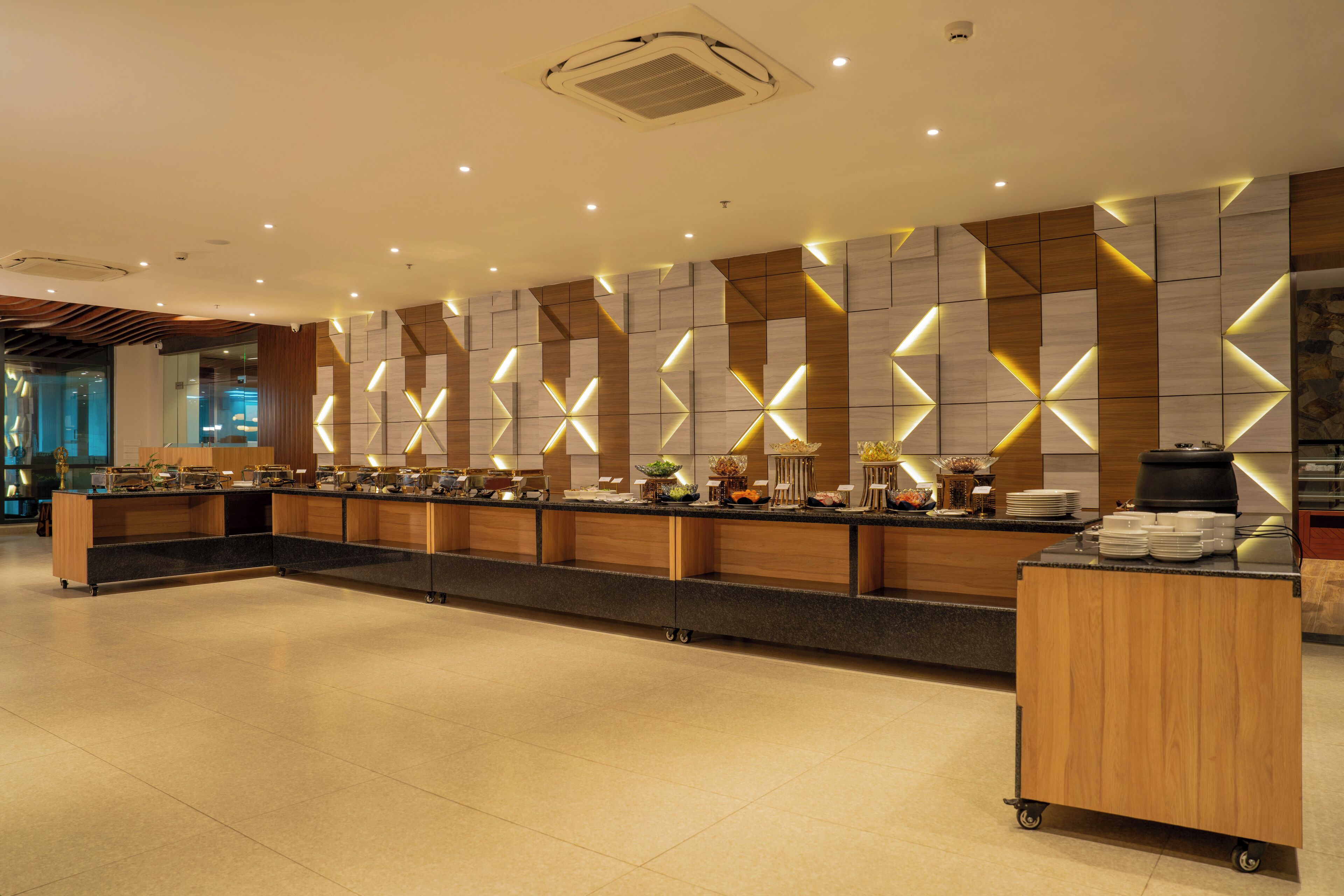 A modern hotel lobby with a long reception desk, a coffee station, and a wall with geometric lighting.