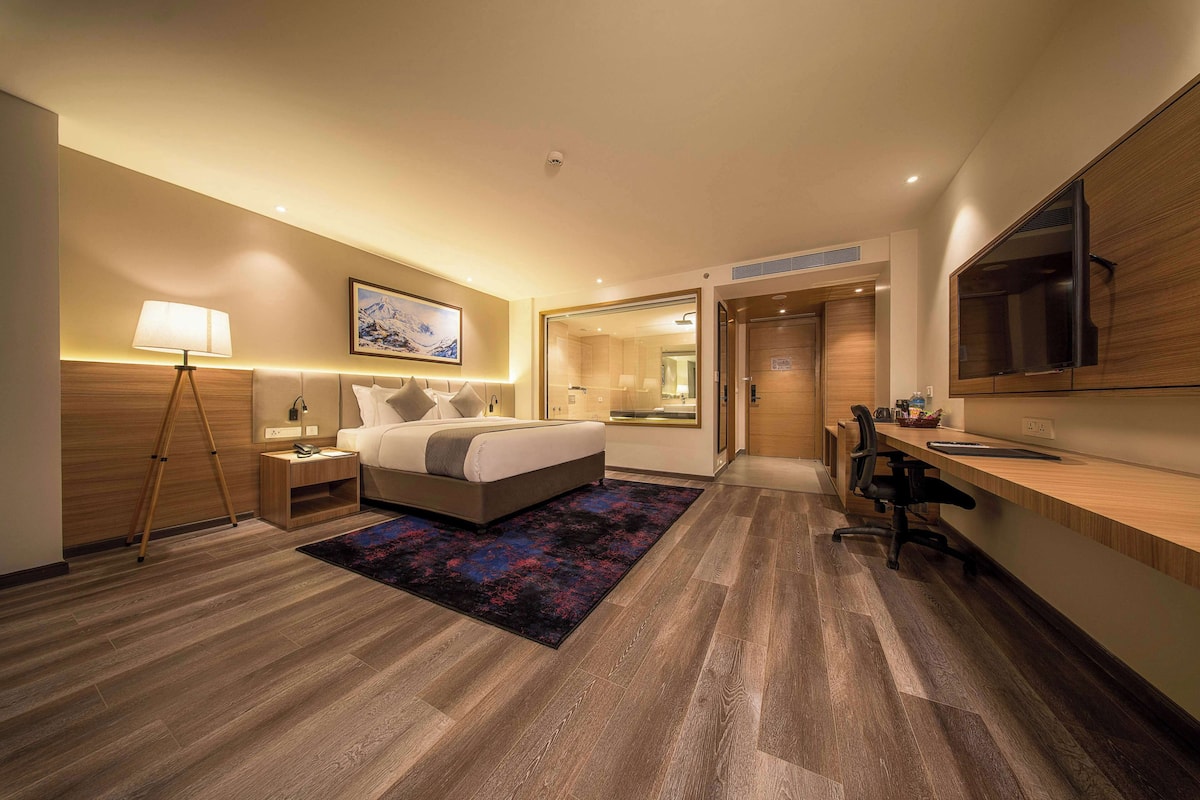 A modern hotel room with a bed, bedside table, lamp, and a desk with a chair.