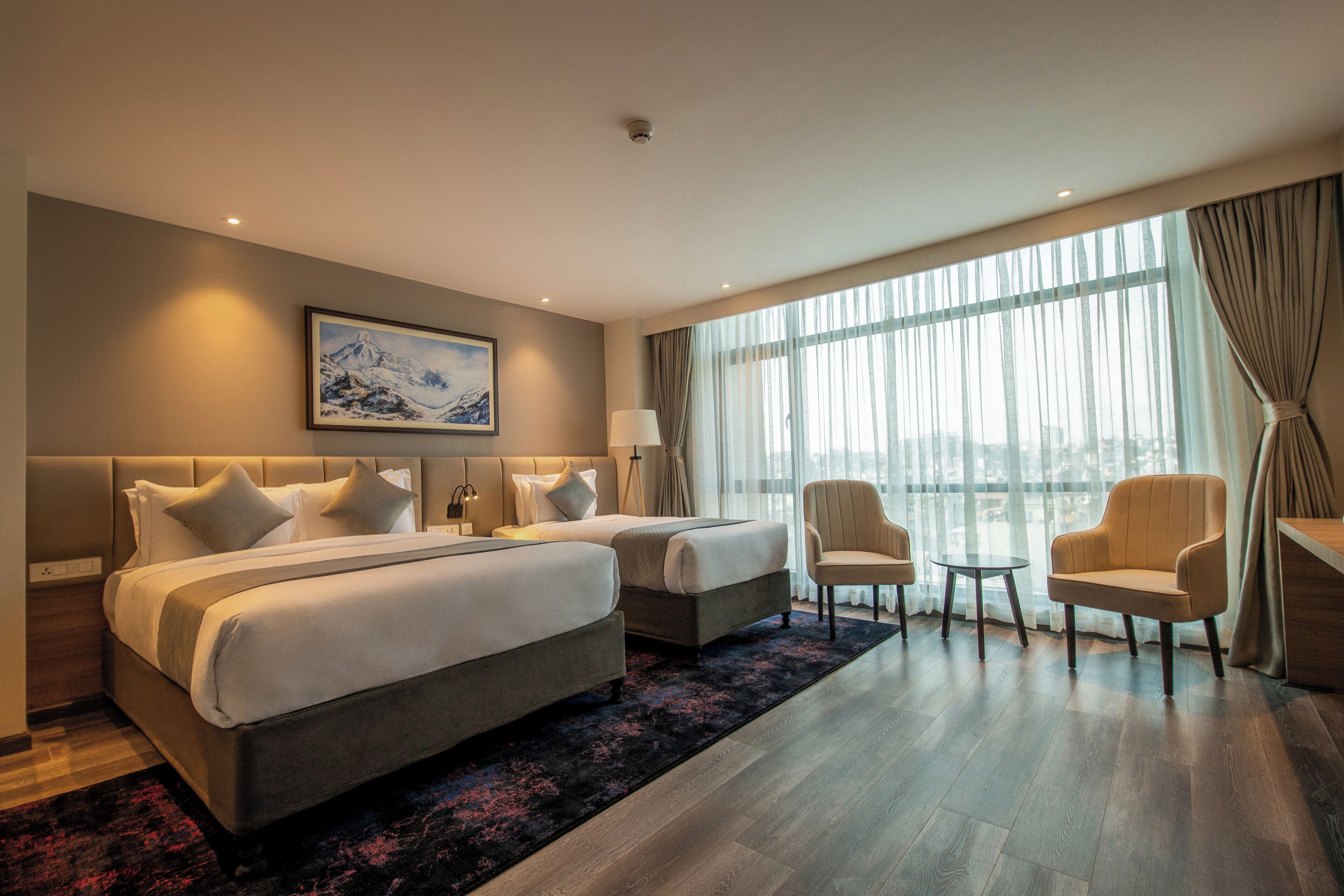 A hotel room with a large bed, two bedside tables, a desk, two chairs, and a large window with curtains.