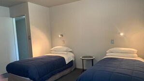 A room with two beds, each with a blue comforter and white pillows. There is a small round table between the beds, and a door leading to another room. - Spires Barn Lodge (Christchurch)