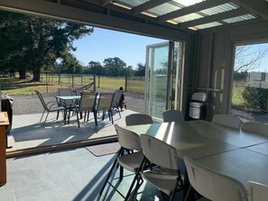 An outdoor patio area with a table and chairs, a glass door leading outside, and a view of a grassy field. - Spires Barn Lodge (Christchurch)