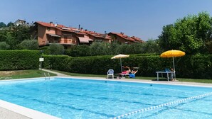 A swimming pool with clear blue water, surrounded by a paved deck. There are lounge chairs and umbrellas, and a building with red roofs in the background. - Appartamento Giada in Padenghe sul Garda (Padenghe sul Garda)