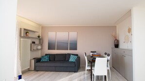 A modern living room with a grey sofa, white chairs, a dining table, and a kitchen area with cabinets and a radiator. - Appartamento Giada in Padenghe sul Garda (Padenghe sul Garda)