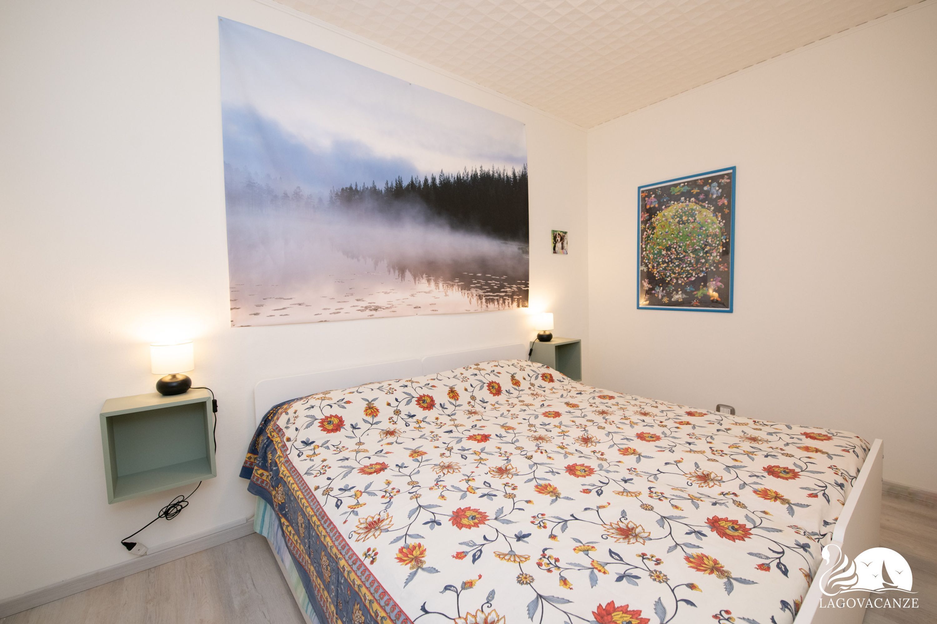A bedroom with a bed, a nightstand, a wall-mounted shelf, a painting, and a framed picture.