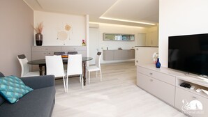 A modern living room with a grey sofa, a dining area with white chairs, a TV on a stand, and a kitchen in the background. - Appartamento Giada in Padenghe sul Garda (Padenghe sul Garda)