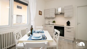 A modern kitchen with white cabinetry, a dining table set for four, and a window with white curtains. - La Torretta - App Catullo (Moniga del Garda)
