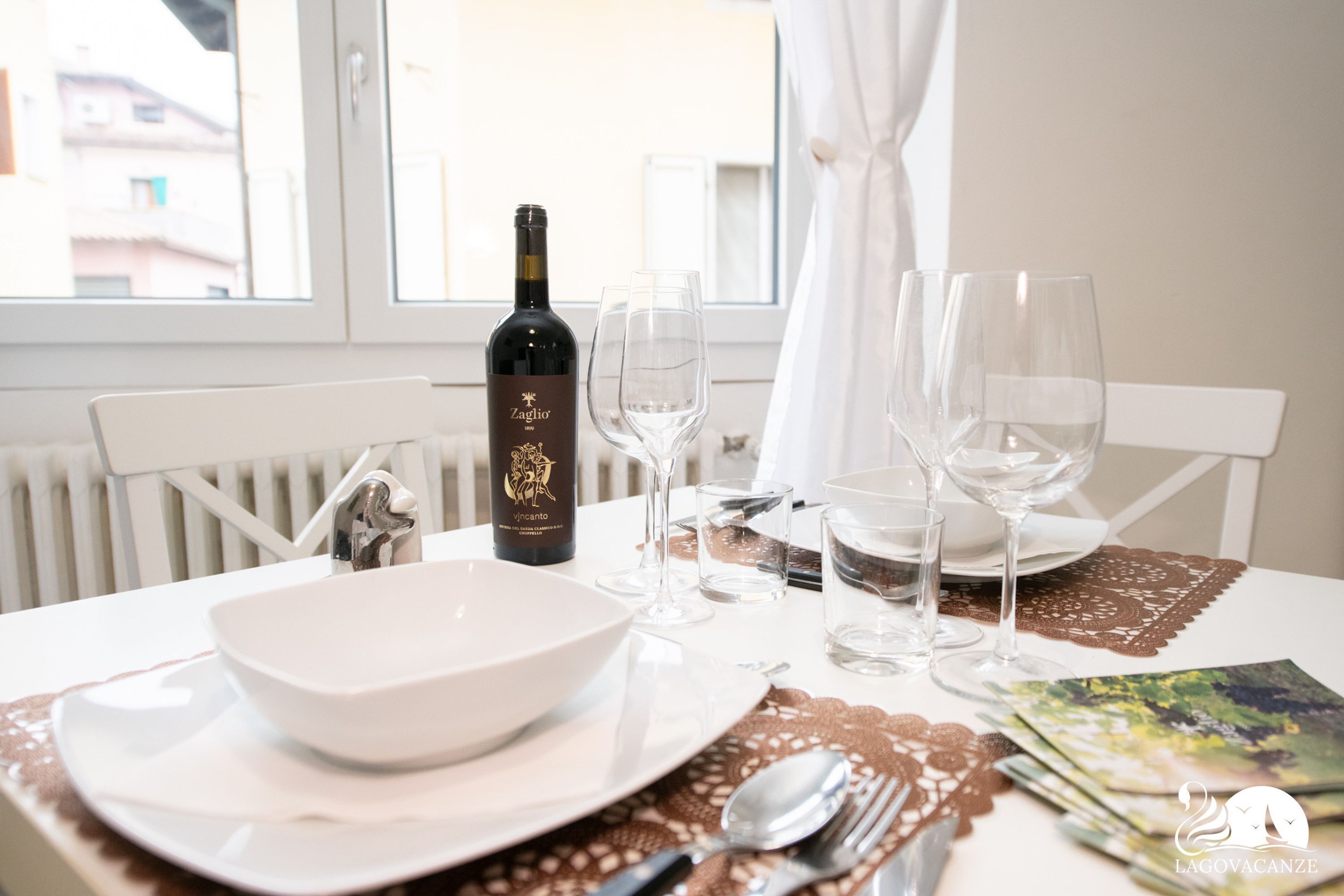 Une table à manger dressée avec un saladier, des assiettes, des couverts et des verres à vin, ainsi qu’une bouteille de vin et une vue sur la fenêtre.
