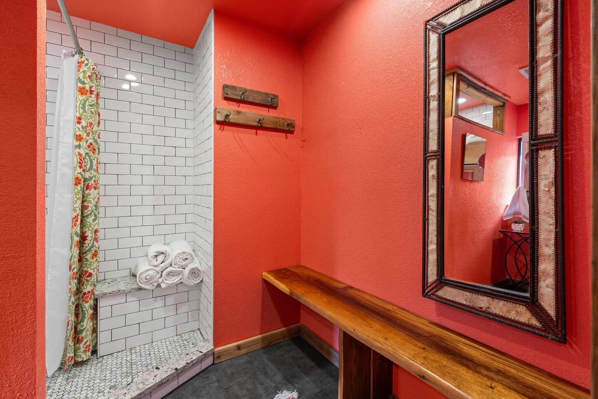 A bathroom with a red wall, a white tiled shower area, a wooden bench, and a mirror with a decorative frame.